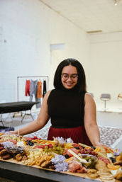 Smiling woman arranging a large colorful charcuterie board with cheese, crackers, grapes and cured meats on a wooden table in a bright white indoor studio event space.