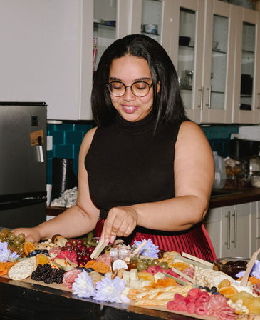 Person wearing glasses and a black sleeveless top arranging a colorful charcuterie board in a home kitchen with cheeses, cured meats, grapes, crackers, dried fruit and edible flowers
