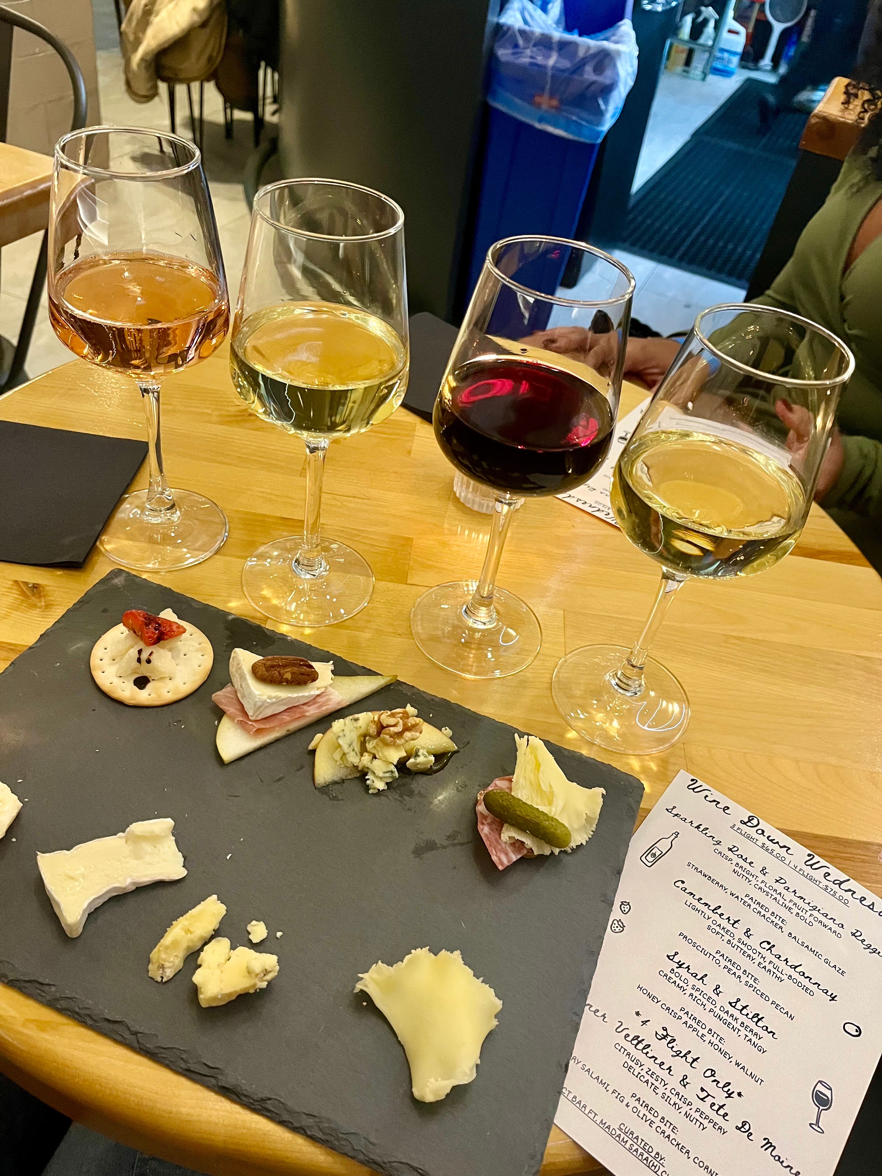 Four-glass wine flight (rosé, two whites, red) with a slate cheese and charcuterie board on a wooden table at a cozy indoor wine bar, menu visible