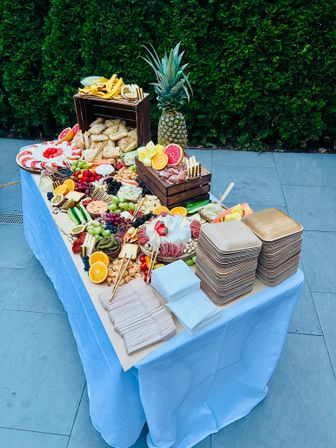 Backyard patio charcuterie spread on a blue‑clothed table with a pineapple centerpiece, wooden crates, stacked eco plates and napkins, assorted cheeses, cured meats, crackers, breads, citrus slices, grapes and berries.