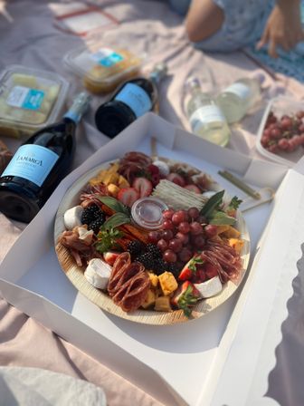 Gourmet charcuterie board with grapes, strawberries, blackberries, cheeses, cured meats and crackers next to bottles of sparkling wine on a picnic blanket