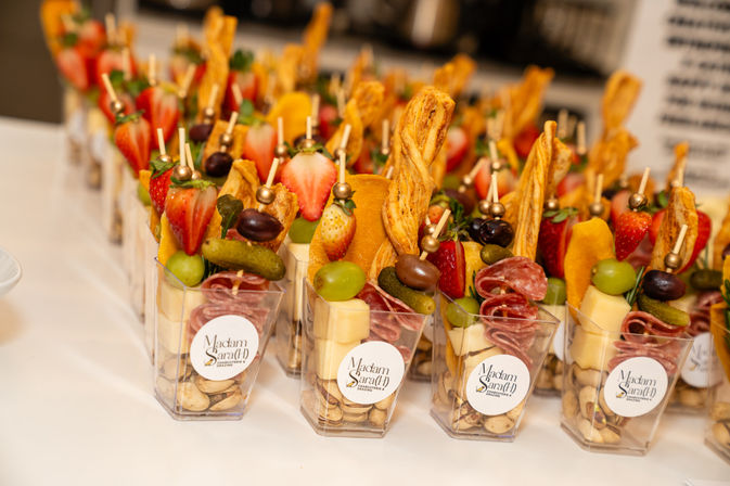 Mini charcuterie cups with skewers of strawberries, grapes, olives, cornichons, cheese cubes, folded salami and twisted breadsticks on a white table at a catered event