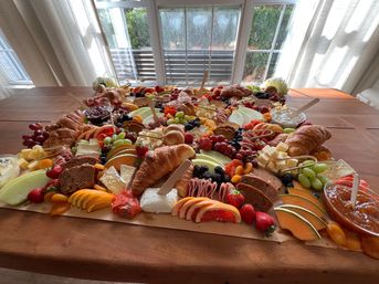 Brunch-ready grazing charcuterie board on a wooden dining table by a sunny window — croissants, assorted cheeses, cured meats, grapes, berries, melon and jams.