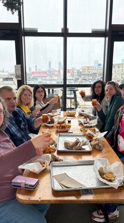 Group of people toasting with small glasses over burgers and fries at a casual rooftop eatery, rain-speckled floor-to-ceiling windows framing a cloudy city skyline.