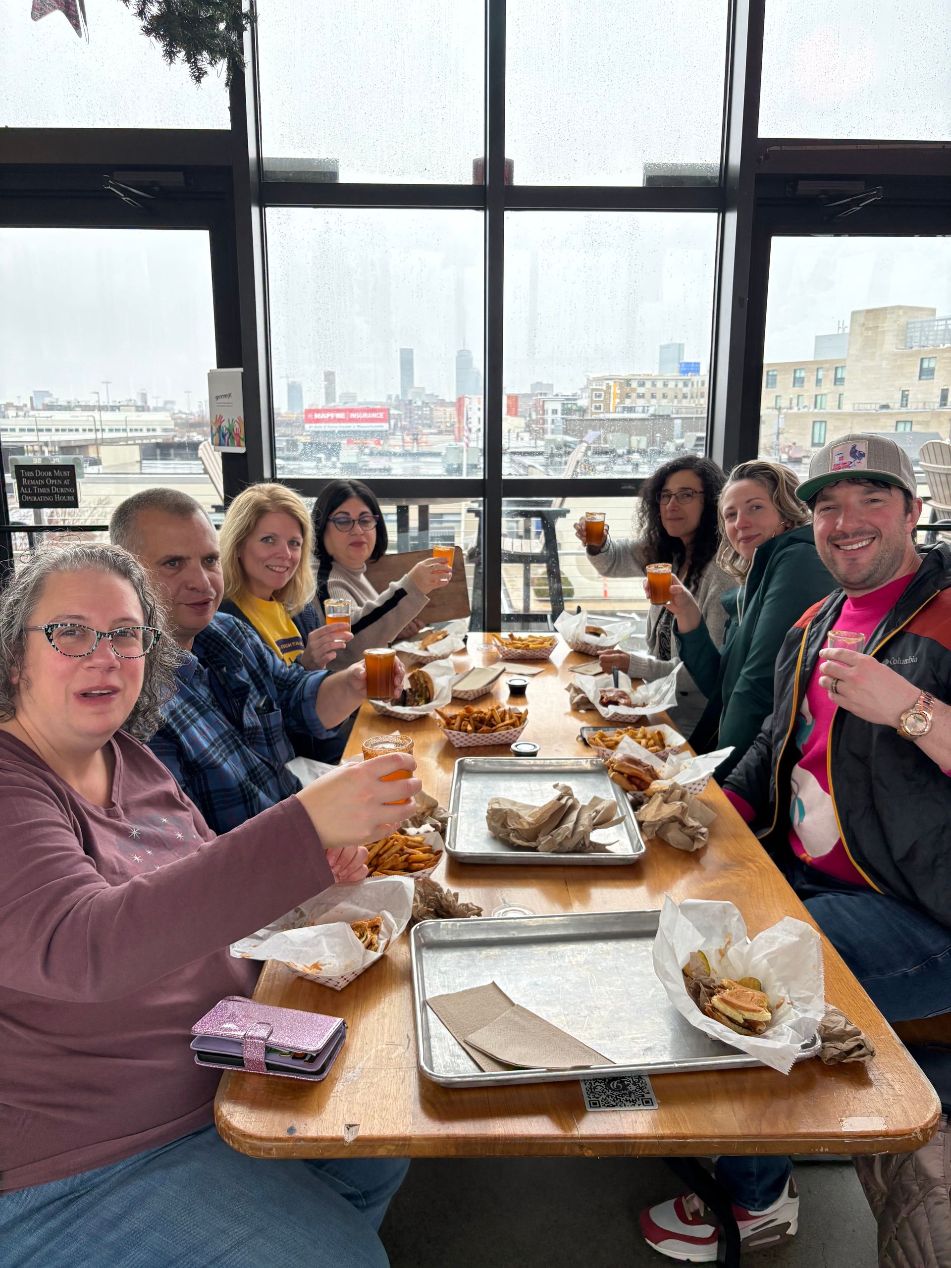 Group of people toasting with small glasses over burgers and fries at a casual rooftop eatery, rain-speckled floor-to-ceiling windows framing a cloudy city skyline.