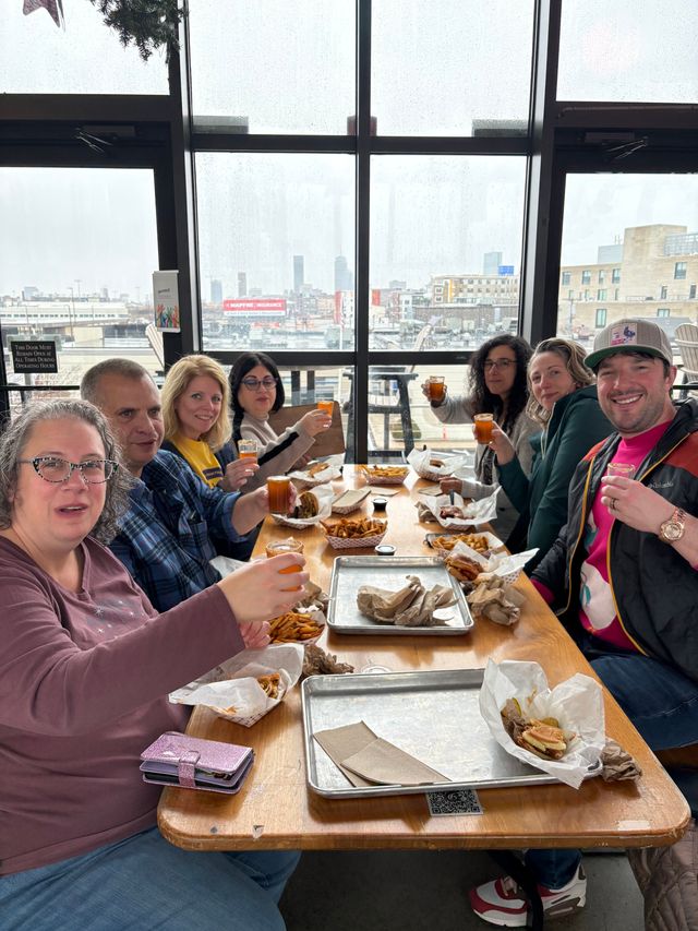 Group of people toasting with small glasses over burgers and fries at a casual rooftop eatery, rain-speckled floor-to-ceiling windows framing a cloudy city skyline.