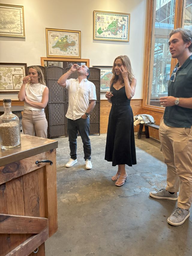 Four adults in a cozy craft distillery tasting room sampling shots at a wooden bar, with framed vintage maps on the wall and stainless-steel brewing tanks visible through a large window.