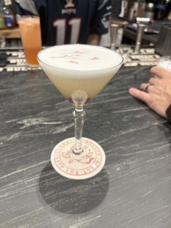 Frothy sour-style cocktail in a martini glass topped with red bitters garnish, resting on a round coaster on a dark bar countertop — craft cocktail at a cocktail bar.