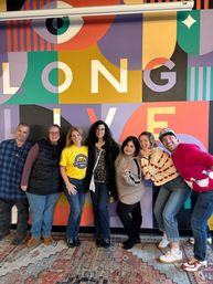 Seven people smiling and posing in front of a bright geometric mural that reads "LONG LIVE" in a modern indoor space — colorful wall, casual group photo.