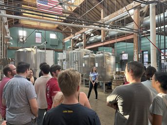 Group on a craft brewery tour inside a rustic industrial building with exposed wooden beams, large stainless-steel fermentation tanks, an American flag overhead, and a guide speaking to the crowd.