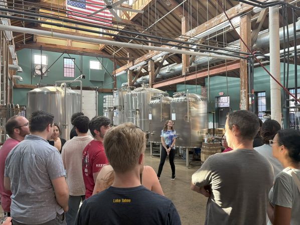 Group on a craft brewery tour inside a rustic industrial building with exposed wooden beams, large stainless-steel fermentation tanks, an American flag overhead, and a guide speaking to the crowd.