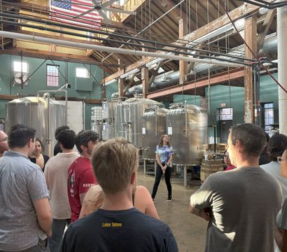 Group on a craft brewery tour inside a rustic industrial building with exposed wooden beams, large stainless-steel fermentation tanks, an American flag overhead, and a guide speaking to the crowd.