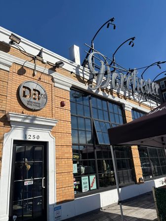 Sunlit brick brewery taproom storefront with large industrial windows, decorative metal rooftop sign, sidewalk canopy and an 'open' neon sign against a clear blue sky.