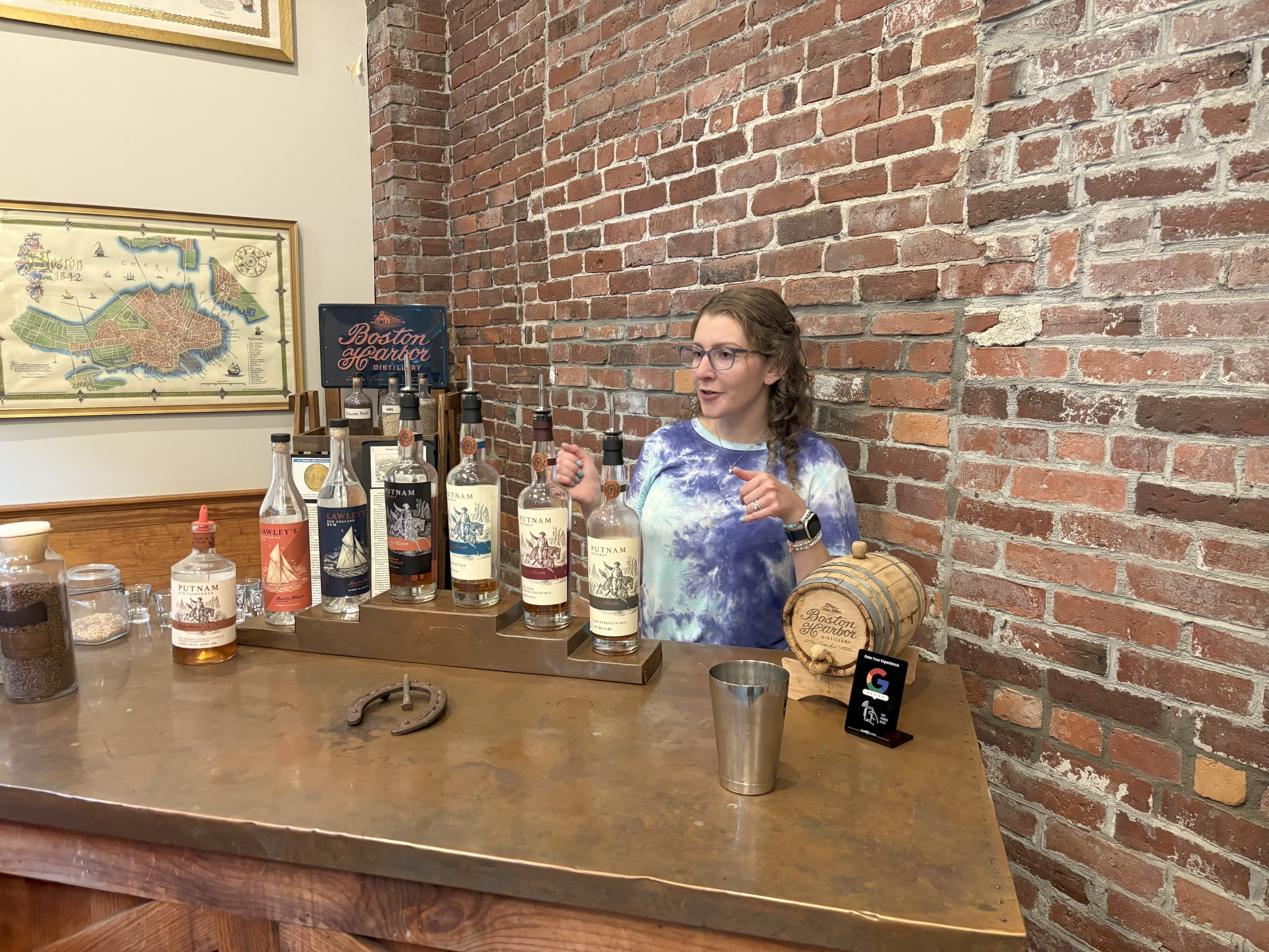 Staff member gesturing while explaining spirits at a distillery tasting bar with lined bottles, a small wooden tasting barrel, metal cup and horseshoe on the counter against an exposed red brick wall and framed map.
