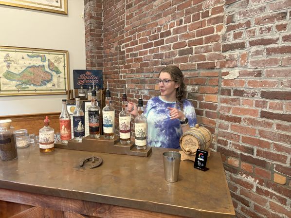 Staff member gesturing while explaining spirits at a distillery tasting bar with lined bottles, a small wooden tasting barrel, metal cup and horseshoe on the counter against an exposed red brick wall and framed map.