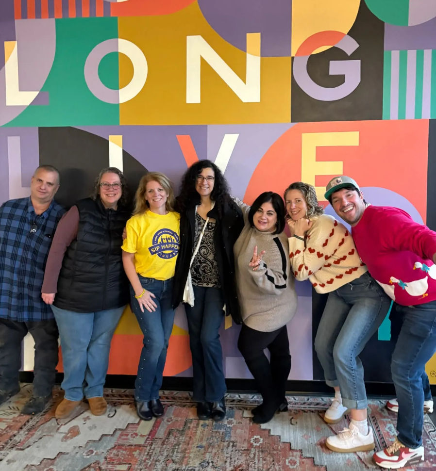 Seven people smiling and posing indoors in casual clothes in front of a colorful geometric mural reading 'LONG LIVE', standing on a patterned rug.