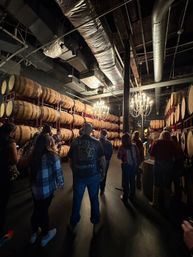 Group on a tasting tour inside an industrial barrel room with rows of stacked oak barrels on red racks, warm chandeliers and exposed ductwork overhead