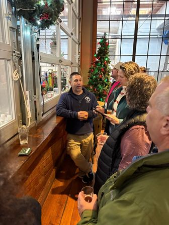 Man chatting with a small group of patrons inside a festive craft brewery taproom with a decorated Christmas tree, wooden bar, industrial windows and glasses of beer.