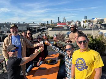 Group of friends toasting with craft beers on a sunny urban rooftop patio, smiling against a city skyline and highway backdrop