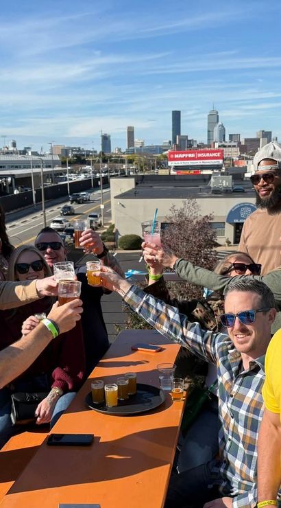 Group of friends toasting with craft beers on a sunny urban rooftop patio, smiling against a city skyline and highway backdrop