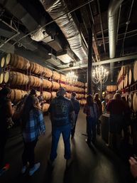 Dimly lit industrial distillery barrel room with stacked oak barrels on racks, exposed ductwork and chandeliers overhead as a group tours a whiskey tasting space.