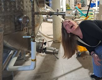 Person laughing and leaning beside a stainless-steel fermentation tank on a brewery production floor with pipes, hoses, and valves.