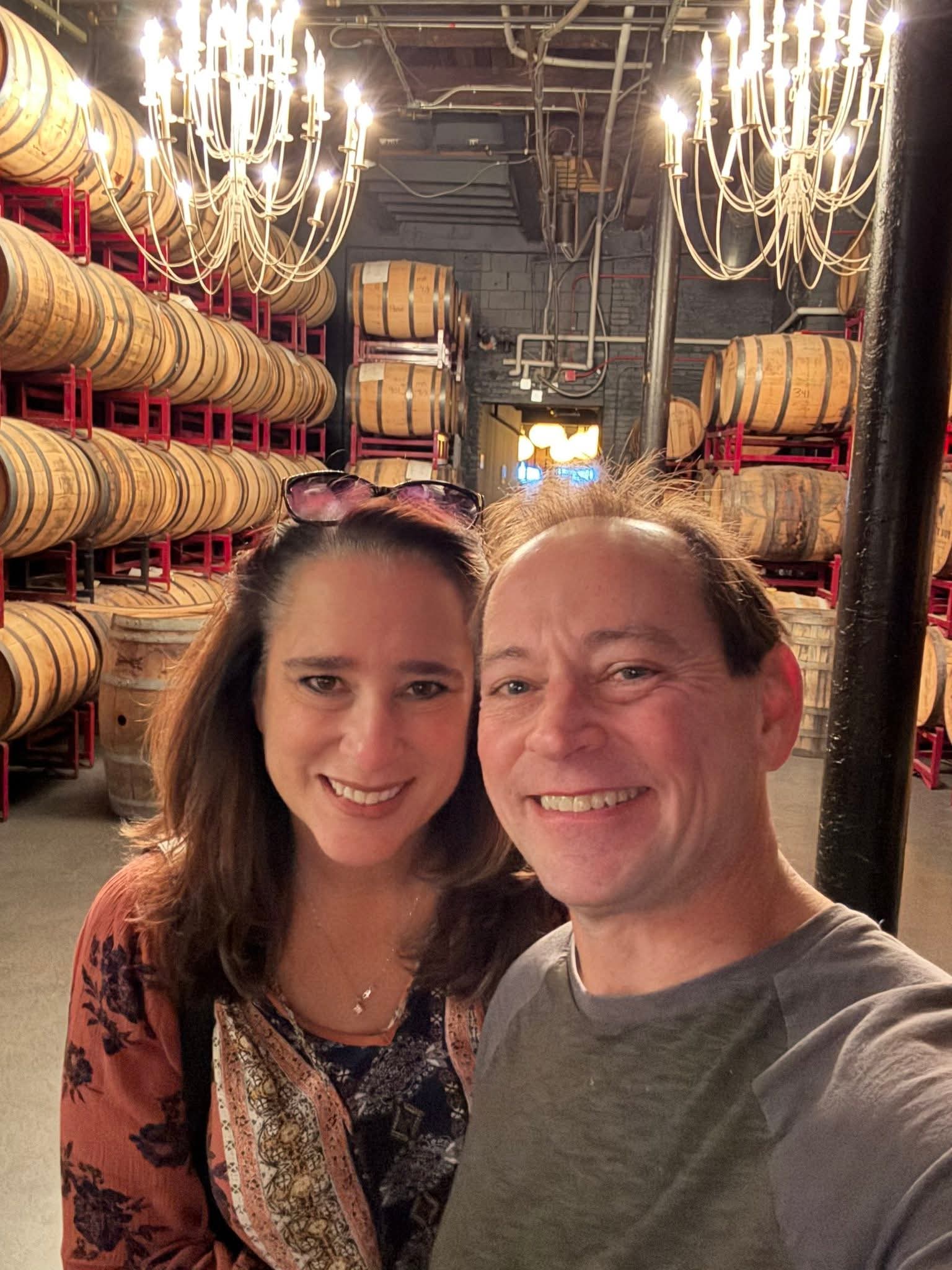 Two smiling adults taking a selfie in a distillery barrel room, framed by rows of stacked oak barrels on red racks and glowing chandeliers overhead.