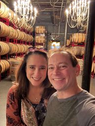 Two smiling adults taking a selfie in a distillery barrel room, framed by rows of stacked oak barrels on red racks and glowing chandeliers overhead.