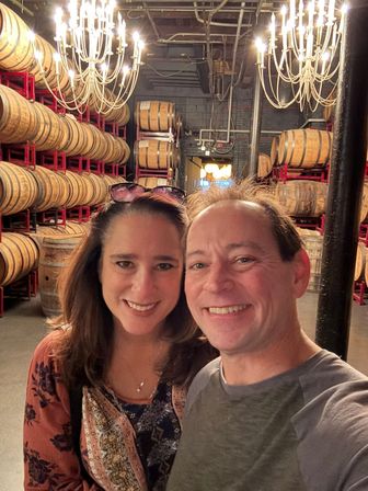 Two smiling adults taking a selfie in a distillery barrel room, framed by rows of stacked oak barrels on red racks and glowing chandeliers overhead.