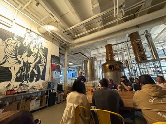 Group seated at a long wooden bar in an industrial-style distillery tasting room with large copper stills, stainless tanks, exposed white ductwork and a bold horse mural on a brick wall.