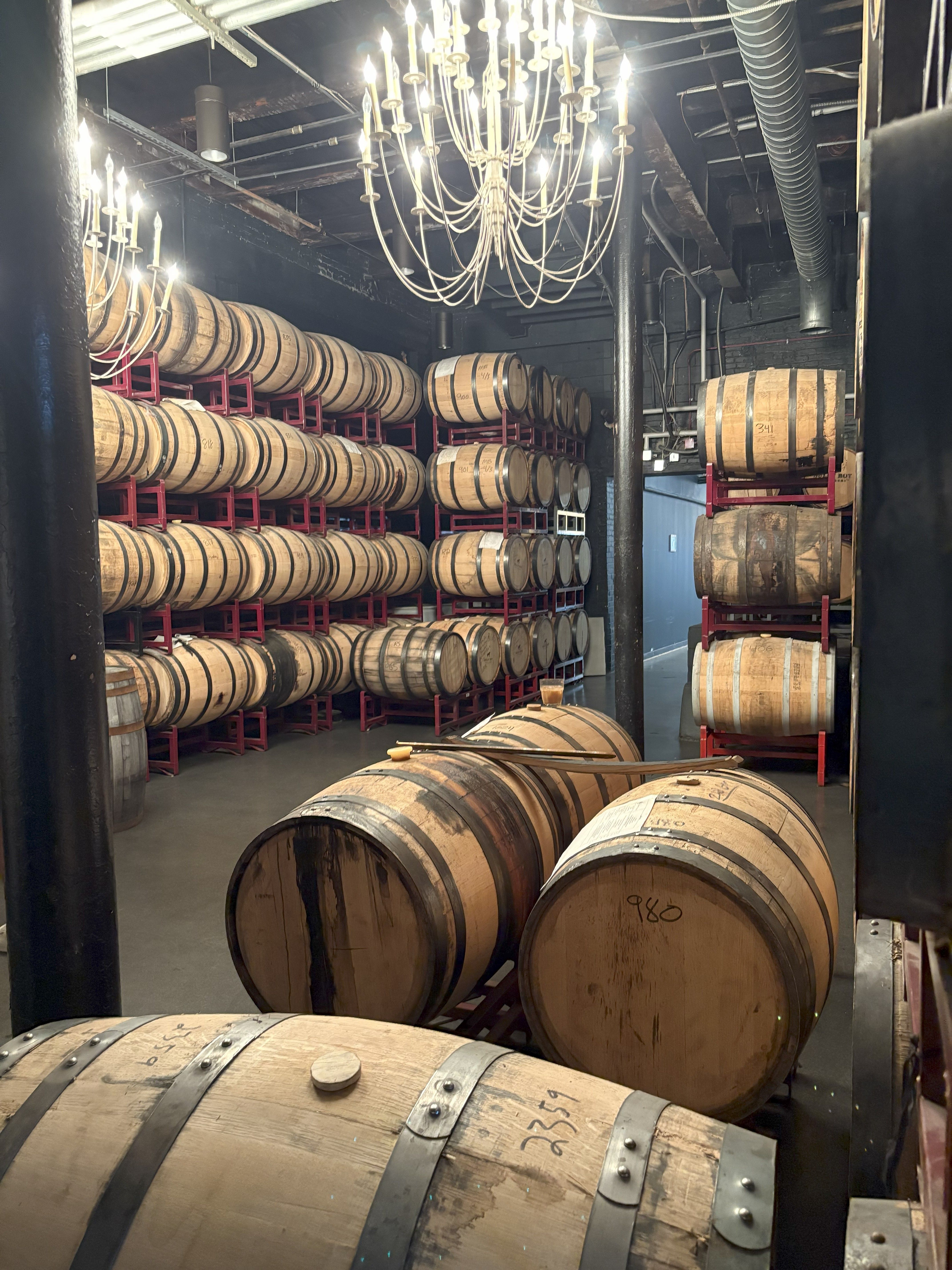 Barrel room with rows of wooden aging barrels stacked on red metal racks under glowing chandeliers in an industrial-style cellar.