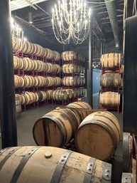 Barrel room with rows of wooden aging barrels stacked on red metal racks under glowing chandeliers in an industrial-style cellar.