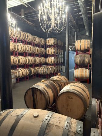 Barrel room with rows of wooden aging barrels stacked on red metal racks under glowing chandeliers in an industrial-style cellar.