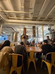 People seated at a long wooden communal table on yellow metal chairs in an industrial-style distillery tasting room with copper column stills, stainless tanks and exposed white ceiling pipes.