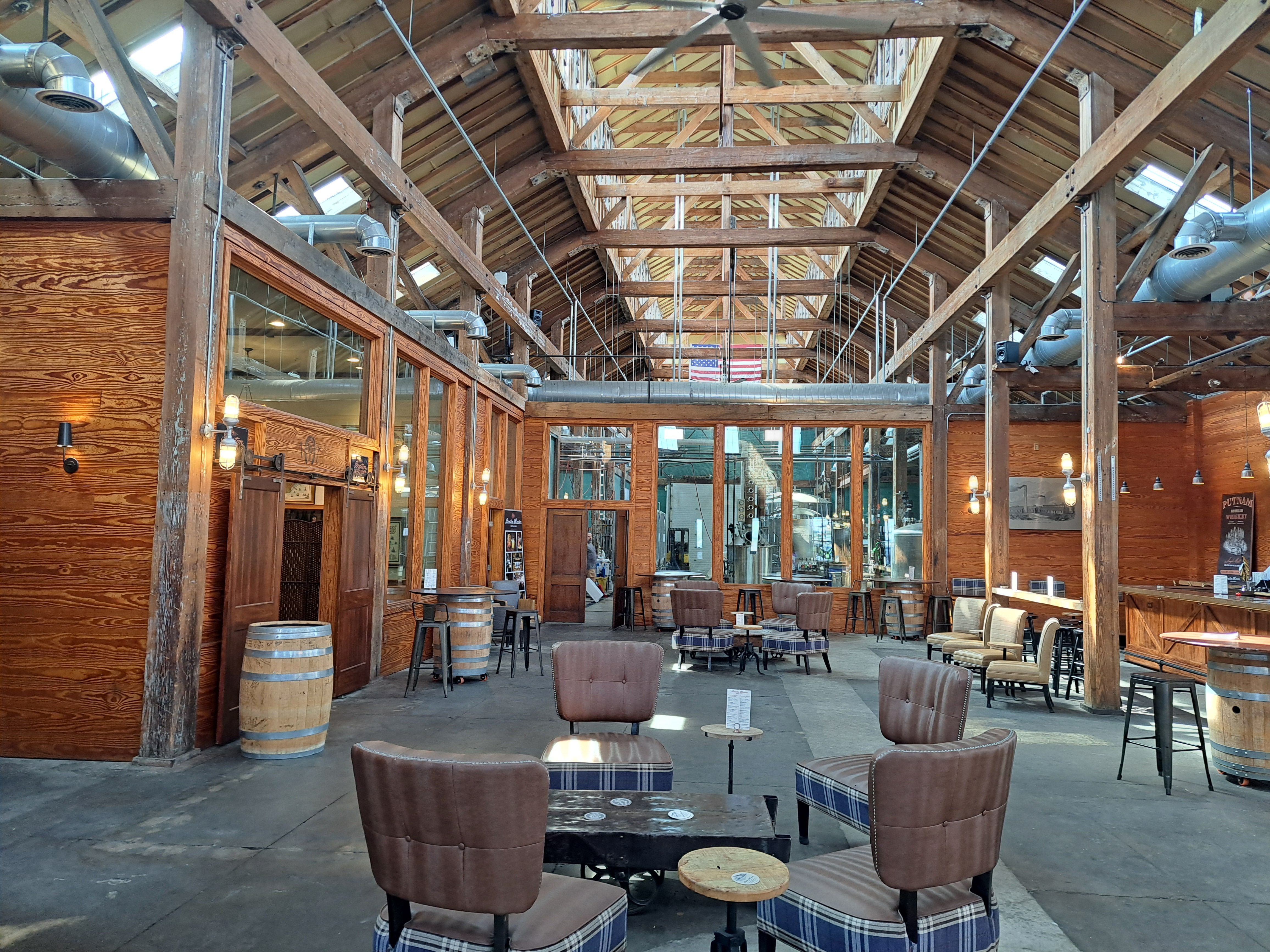 Sunlit industrial-style tasting room in a converted wooden warehouse with exposed beams, skylights, ductwork, barrel tables, and cozy leather and plaid seating.