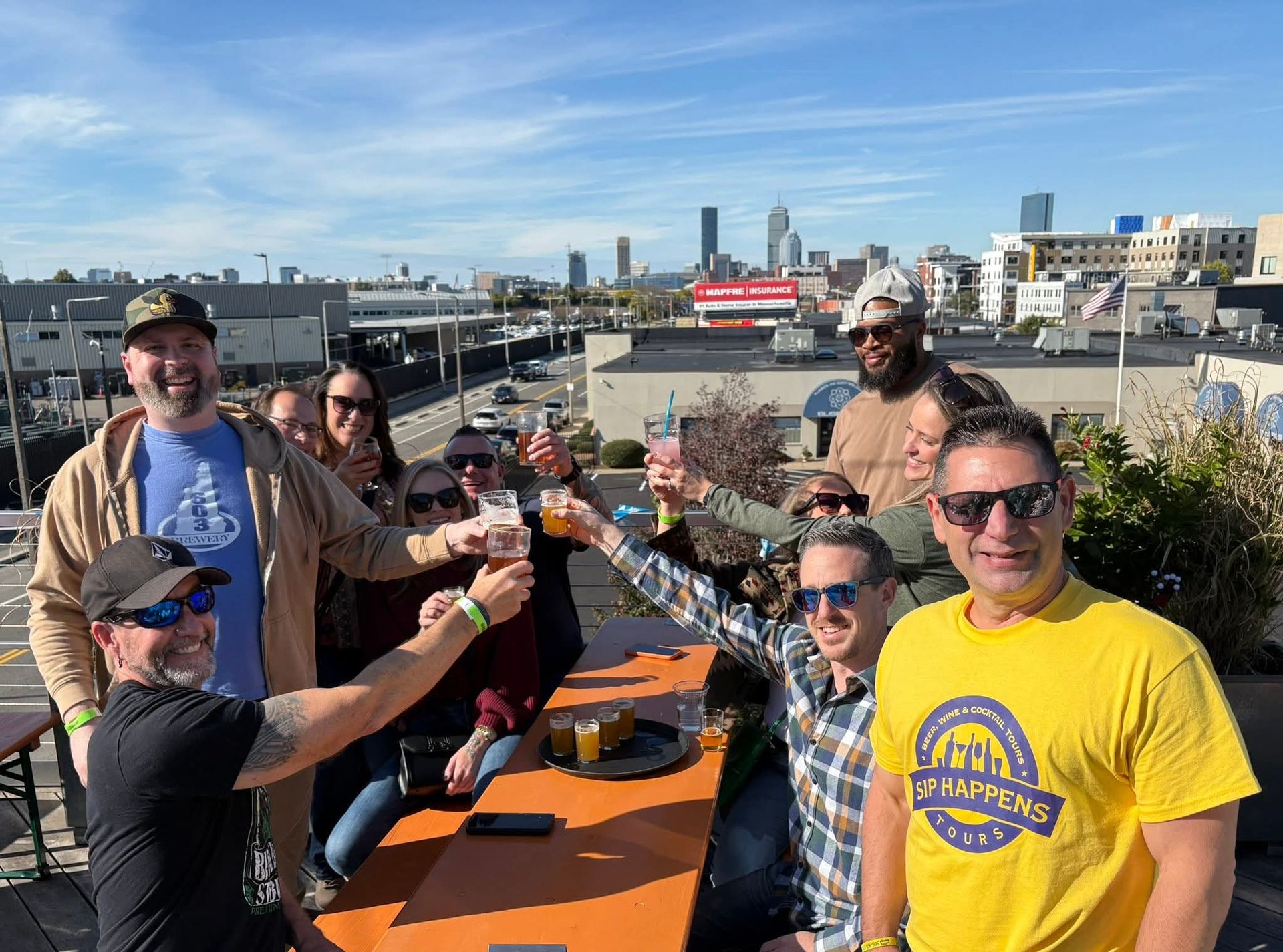 Cheerful group toasting with beers and cocktails on a sunny rooftop patio, tasting flights on the table and a downtown city skyline in the background.