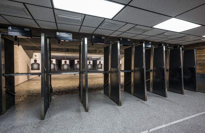 Indoor shooting range with numbered lanes and black partitions, paper silhouette bullseye targets at the far back, spent shell casings on the floor and bright drop-ceiling lights overhead.