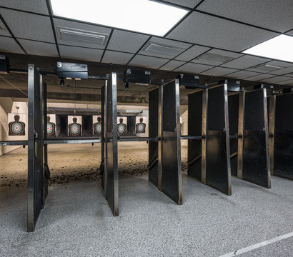 Indoor shooting range with numbered lanes and black partitions, paper silhouette bullseye targets at the far back, spent shell casings on the floor and bright drop-ceiling lights overhead.