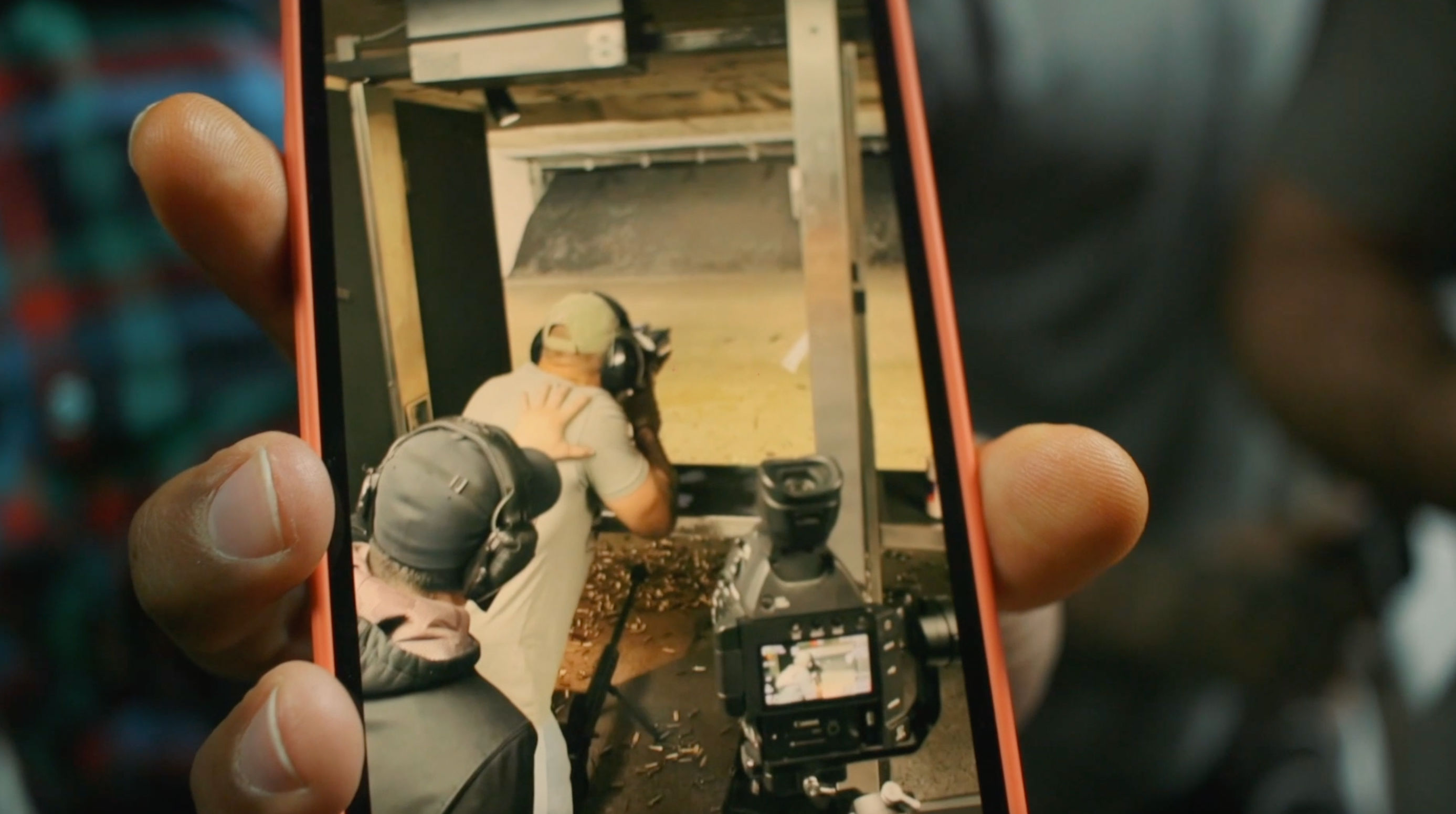 Close-up smartphone screen showing two people with ear protection at an indoor shooting range — one guiding the other, a camera on a tripod and brass casings on the floor.
