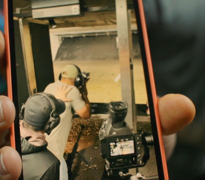 Close-up smartphone screen showing two people with ear protection at an indoor shooting range — one guiding the other, a camera on a tripod and brass casings on the floor.
