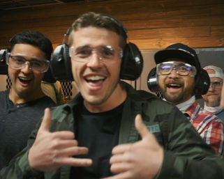 Cheerful group of four men wearing ear protection and safety glasses, posing and making hand gestures inside an indoor shooting range with wood-paneled walls.