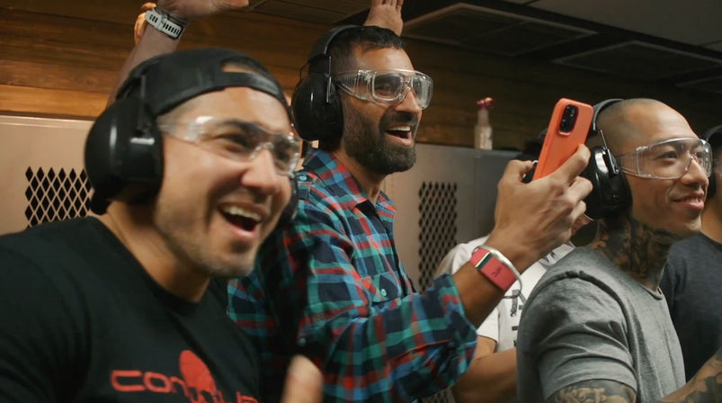 Three men at an indoor shooting range wearing ear protection and safety goggles, smiling and cheering while one records with a smartphone.