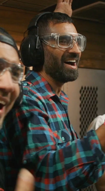 Three men at an indoor shooting range wearing ear protection and safety goggles, smiling and cheering while one records with a smartphone.