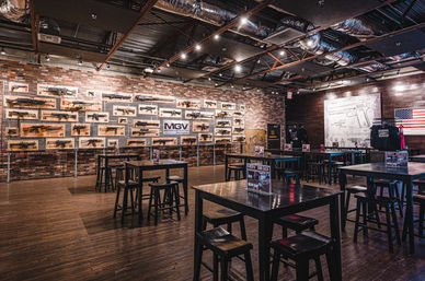 Modern firearms showroom and indoor shooting range lounge with rifles and handguns mounted on a brick wall, wooden high-top tables and stools, exposed industrial ceiling and American flag.