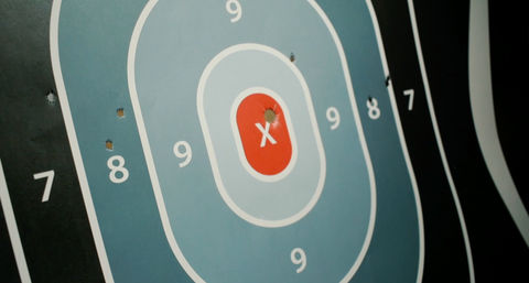 Blue-and-black paper shooting target with a red X bullseye, numbered 7–9 scoring rings, and several bullet holes clustered near the center and in the 8 ring.