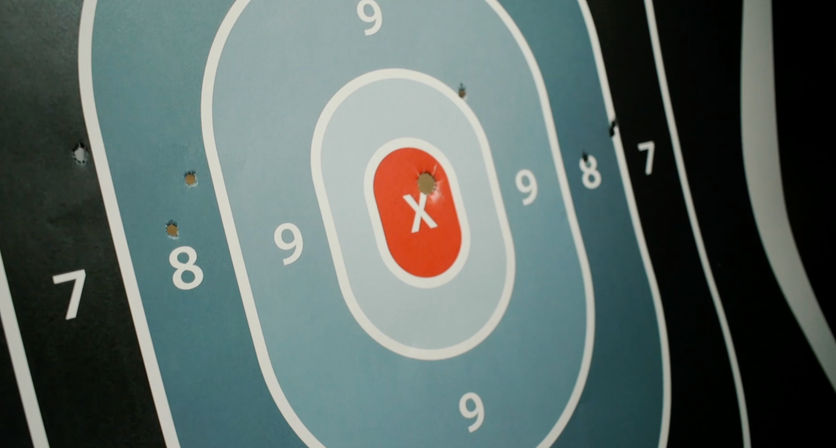 Blue-and-black paper shooting target with a red X bullseye, numbered 7–9 scoring rings, and several bullet holes clustered near the center and in the 8 ring.