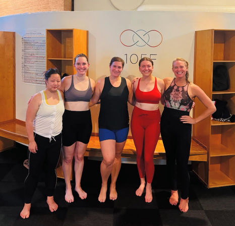 Five women in colorful activewear smiling barefoot in a yoga/fitness studio locker area after class, posing in front of wooden cubbies and a studio wall.