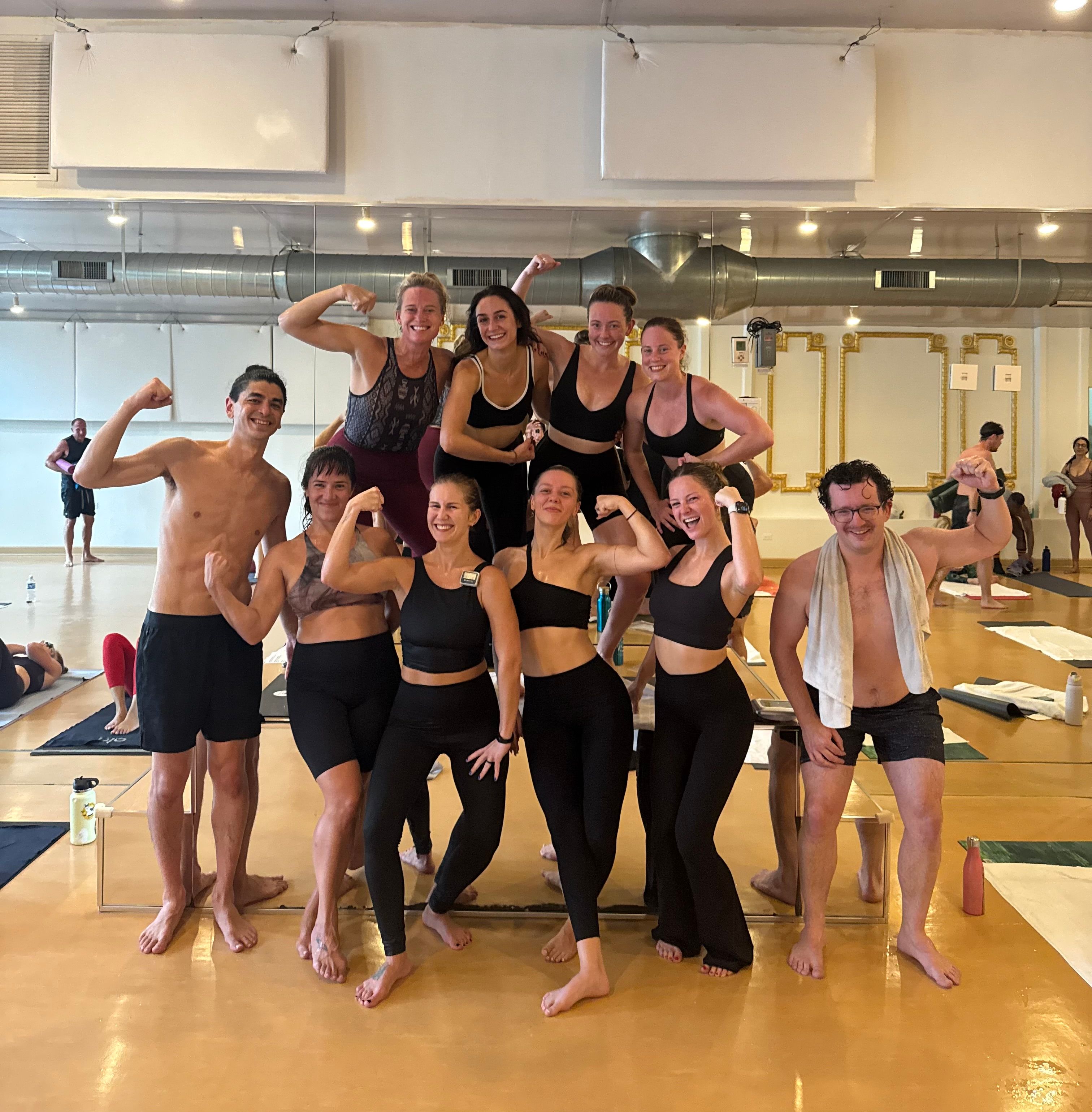 Smiling group of adults in activewear flexing muscles after a sweaty group fitness/yoga class in a mirrored studio with exposed ductwork and hardwood floor