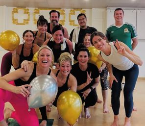 Smiling adults in a bright indoor fitness studio posing after a group workout, holding gold and silver balloons and flashing peace signs — celebratory group fitness class photo.