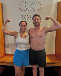 Smiling pair flexing biceps after a workout — woman in a white tank and blue shorts and a shirtless man in black shorts posing in front of a wall with a circular logo and the number 105 in a fitness studio.